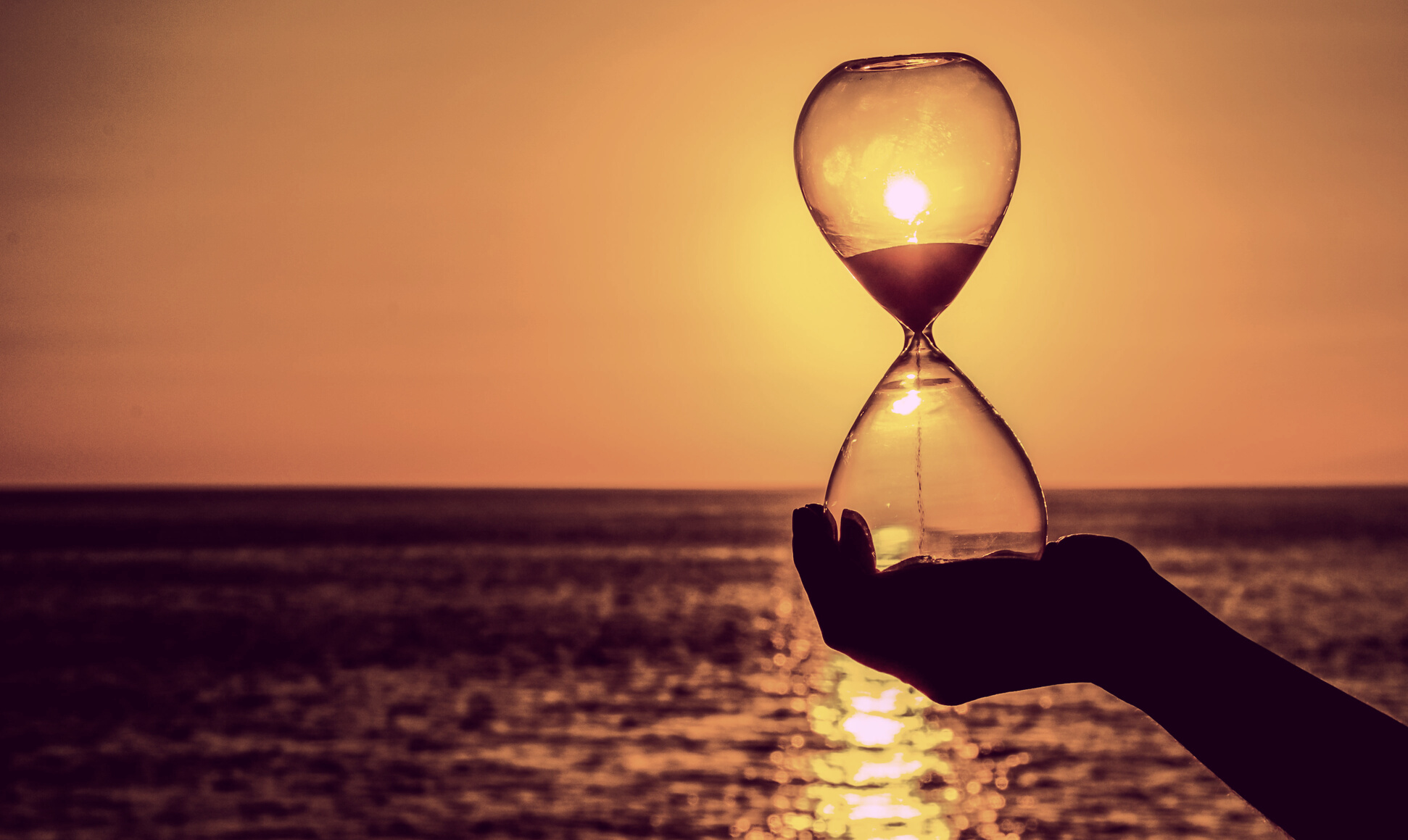 Eine Hand hält eine Sanduhr vor dem Sonnenuntergang am Meer